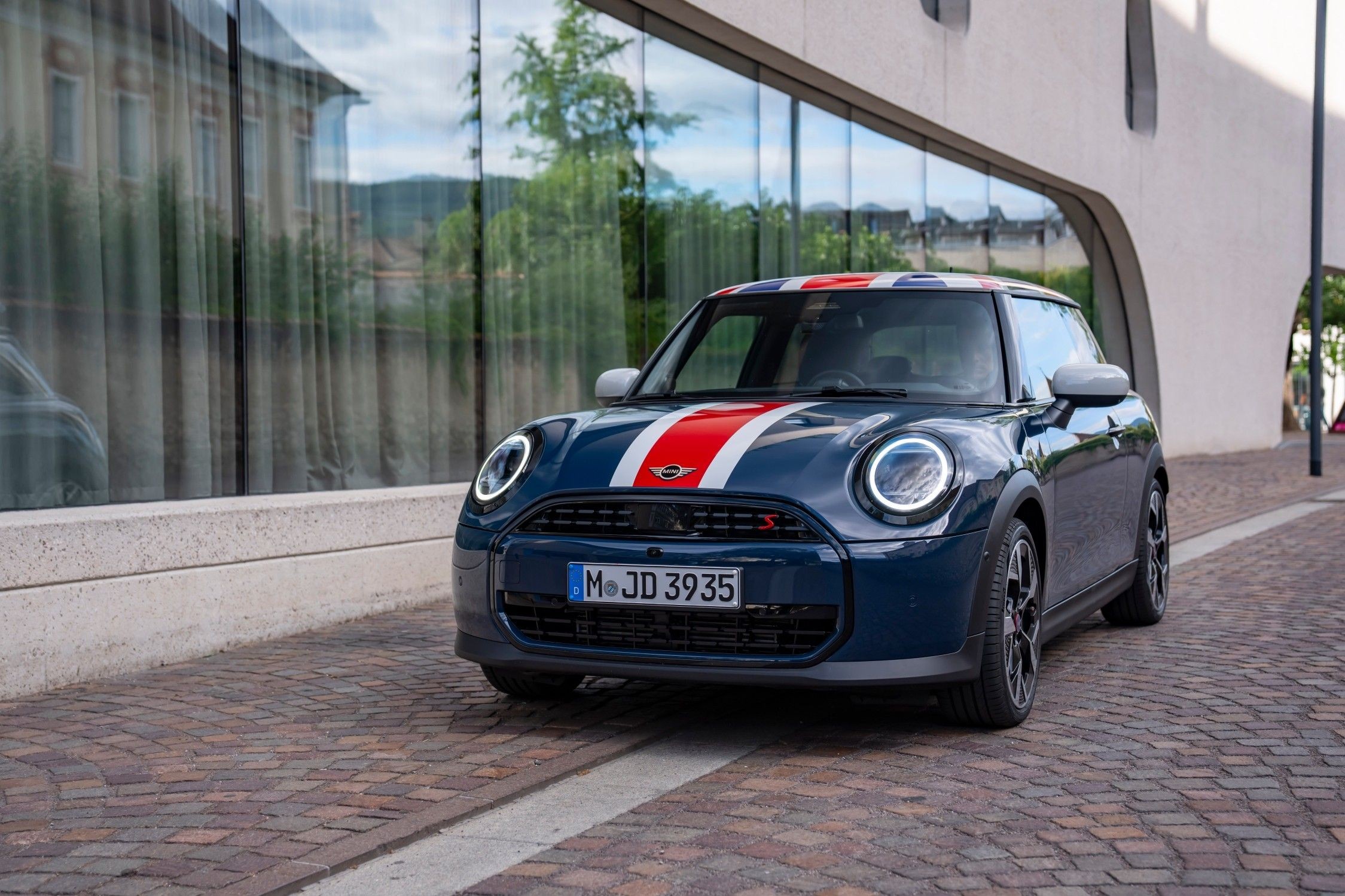 MINI Cooper Oxford Edition azul, com tejadilho Union Jack e faixas vermelhas e brancas no capô, estacionado junto a um edifício moderno